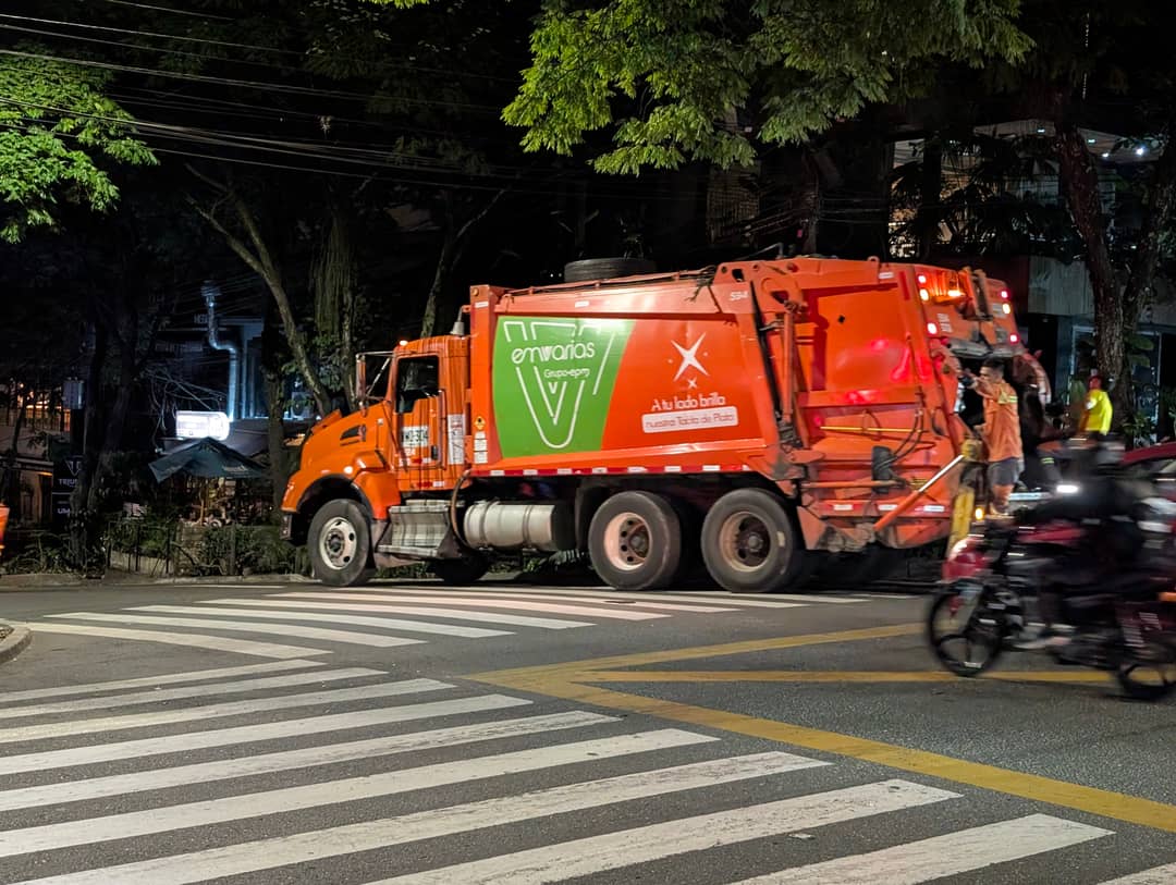 A bin lorry drives down a street full of people. Two men jog alongside, one rides on the back, motorbikes in between.